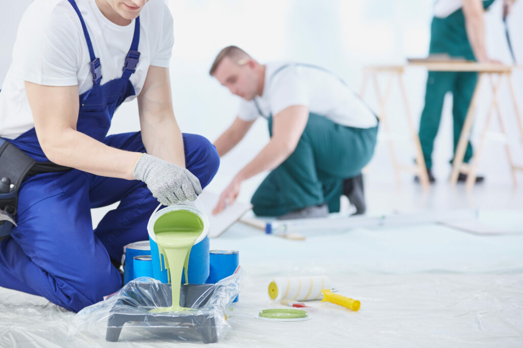 Zwei Maler bereiten sich auf Malerarbeiten vor; einer gießt grüne Farbe in eine Schale, während ein anderer im Hintergrund arbeitet. Farbvorräte sind auf einem mit Plastik bedeckten Boden verstreut. - Lauer maler & raumdesign - Bodenbeläge, Vinylboden, Farben, Sonnenschutz, Malerarbeiten - Bahnhofstr. 5a - 36280 Oberaula